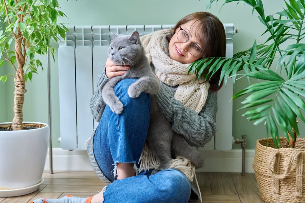A woman in a knitted sweater holding her cat on her lap beside a radiator